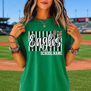 May include: A green short-sleeved t-shirt featuring a graphic design with vertical black and white stripes, the word Eagles in white script, a leopard print bow, and the text SCHOOL NAME in white block letters centered on the chest.