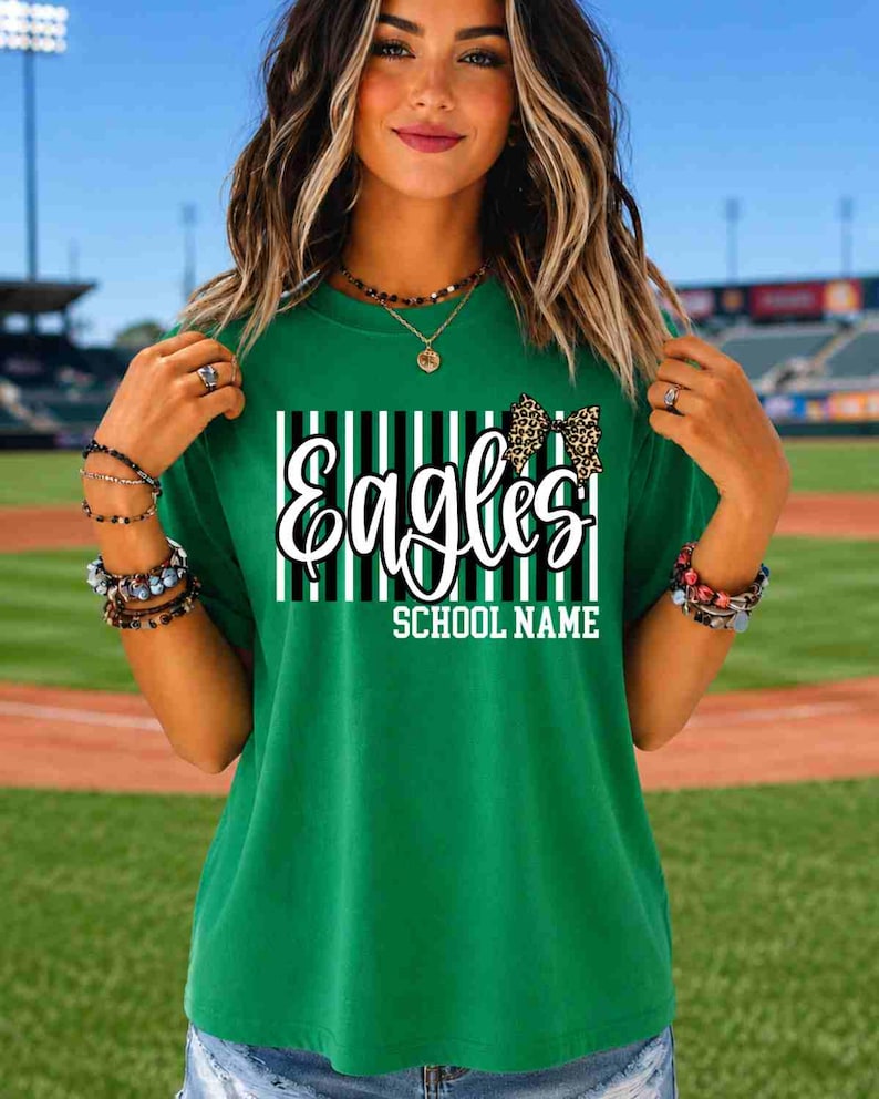 May include: A green short-sleeved t-shirt featuring a graphic design with vertical black and white stripes, the word Eagles in white script, a leopard print bow, and the text SCHOOL NAME in white block letters centered on the chest.