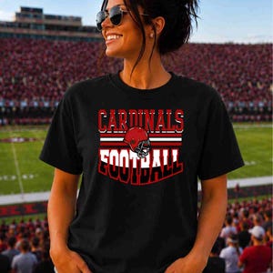 May include: A black short-sleeved t-shirt featuring a graphic design with the text CARDINALS in red and FOOTBALL in white, centered above and below a red football helmet illustration with white and red horizontal stripes.