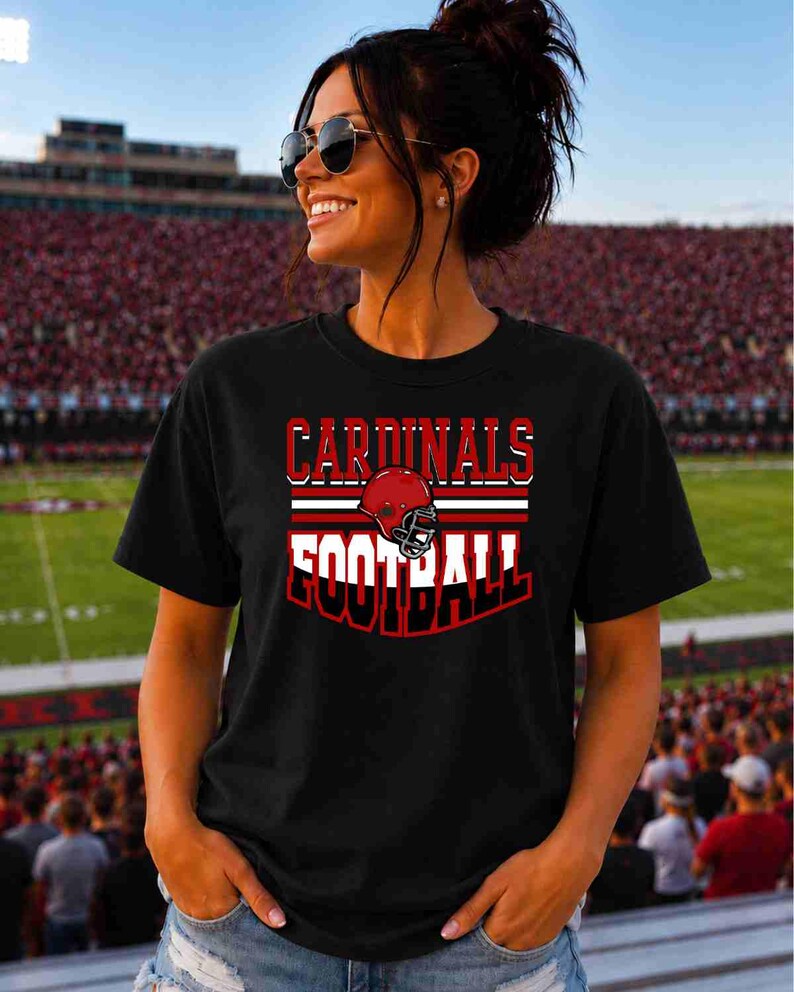 May include: A black short-sleeved t-shirt featuring a graphic design with the text CARDINALS in red and FOOTBALL in white, centered above and below a red football helmet illustration with white and red horizontal stripes.