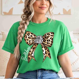 Puede incluir: Una camiseta verde brillante de manga corta con un gr&aacute;fico grande de un lazo con estampado de leopardo centrado en el pecho. En el medio del lazo hay una peque&ntilde;a ilustraci&oacute;n de una cabeza de bulldog gris. La palabra Bulldogs est&aacute; escrita en cursiva blanca.