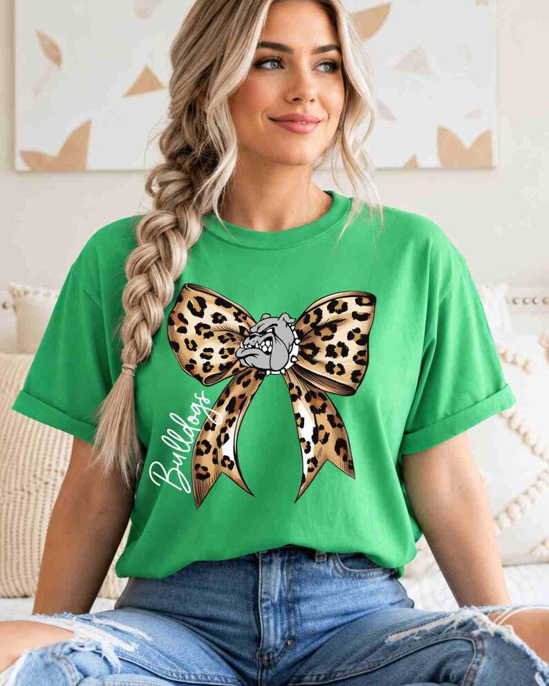 Puede incluir: Una camiseta verde brillante de manga corta con un gr&aacute;fico grande de un lazo con estampado de leopardo centrado en el pecho. En el medio del lazo hay una peque&ntilde;a ilustraci&oacute;n de una cabeza de bulldog gris. La palabra Bulldogs est&aacute; escrita en cursiva blanca.