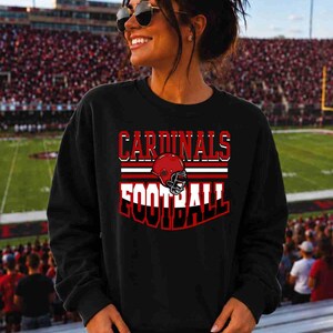 May include: A black long-sleeve sweatshirt featuring a graphic print with the text CARDINALS FOOTBALL in white and red lettering, centered above a red football helmet illustration with a black face mask and horizontal red and white stripes.