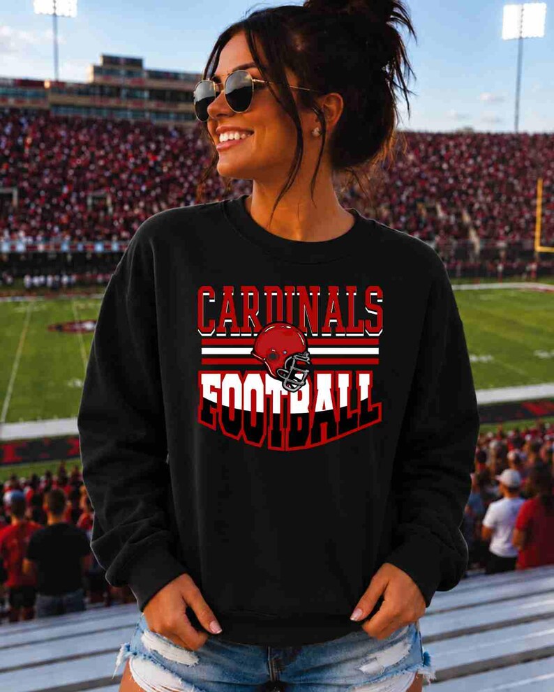 May include: A black long-sleeve sweatshirt featuring a graphic print with the text CARDINALS FOOTBALL in white and red lettering, centered above a red football helmet illustration with a black face mask and horizontal red and white stripes.