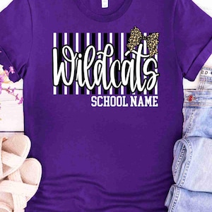 May include: A purple short-sleeved t-shirt featuring a graphic design with vertical black and white stripes. The text "Wildcats" is printed in a white script font, accompanied by a leopard print bow and the words "SCHOOL NAME" in white.