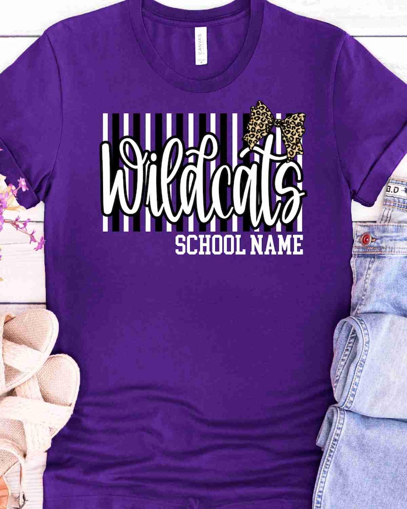 May include: A purple short-sleeved t-shirt featuring a graphic design with vertical black and white stripes. The text "Wildcats" is printed in a white script font, accompanied by a leopard print bow and the words "SCHOOL NAME" in white.