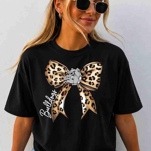 Puede incluir: Una camiseta negra de manga corta con un gr&aacute;fico grande de un lazo con estampado de leopardo centrado en el pecho. Una peque&ntilde;a cabeza de bulldog gris est&aacute; situada en el centro del lazo y la palabra Bulldogs est&aacute; escrita en cursiva blanca.