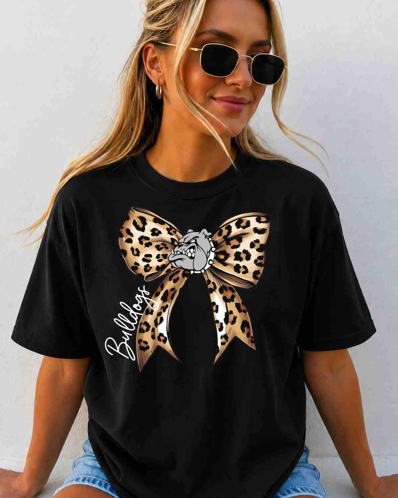 Puede incluir: Una camiseta negra de manga corta con un gr&aacute;fico grande de un lazo con estampado de leopardo centrado en el pecho. Una peque&ntilde;a cabeza de bulldog gris est&aacute; situada en el centro del lazo y la palabra Bulldogs est&aacute; escrita en cursiva blanca.