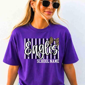 May include: A purple short-sleeve t-shirt featuring a graphic design with vertical black and white stripes, the word "Eagles" in white script, a leopard print bow, and the text "SCHOOL NAME" in white block letters centered on the chest.