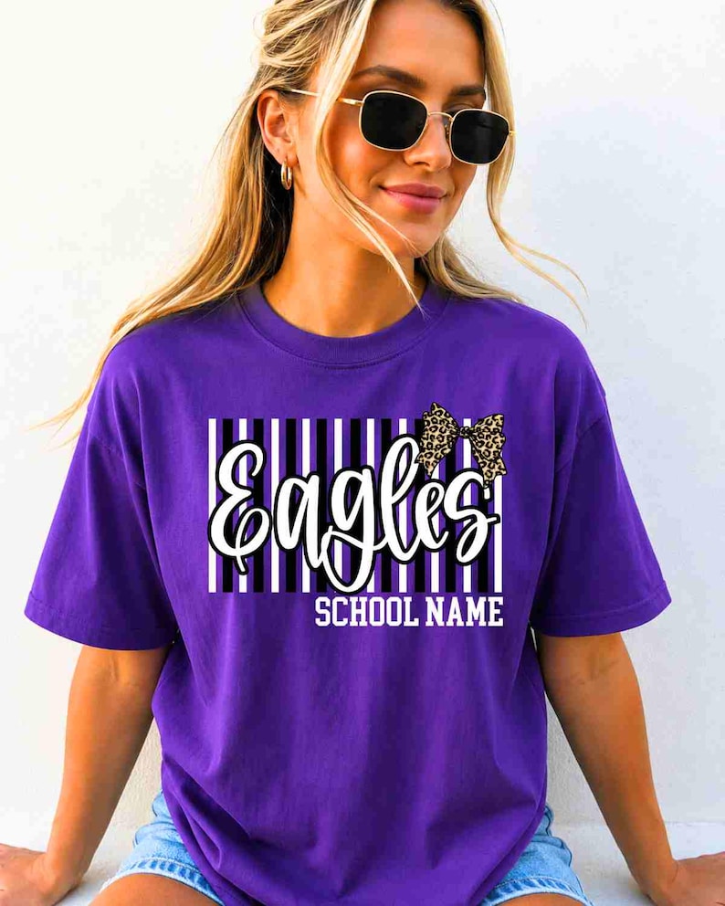 May include: A purple short-sleeve t-shirt featuring a graphic design with vertical black and white stripes, the word "Eagles" in white script, a leopard print bow, and the text "SCHOOL NAME" in white block letters centered on the chest.