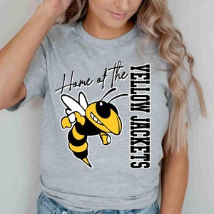 May include: A light gray short-sleeved t-shirt featuring a graphic of a yellow and black hornet. The text "Home of the" is written in a black script font above the hornet, and the words "YELLOW JACKETS" are printed vertically in black block letters.