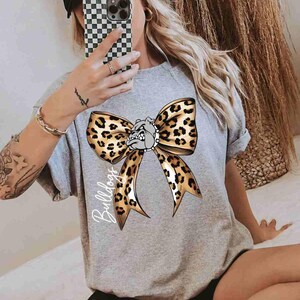 Puede incluir: Una camiseta gris jaspeado de manga corta con un gran estampado gr&aacute;fico de un lazo con estampado de leopardo centrado en el pecho. El lazo incluye una peque&ntilde;a ilustraci&oacute;n de una cara de bulldog en el centro y la palabra Bulldogs escrita en blanco.