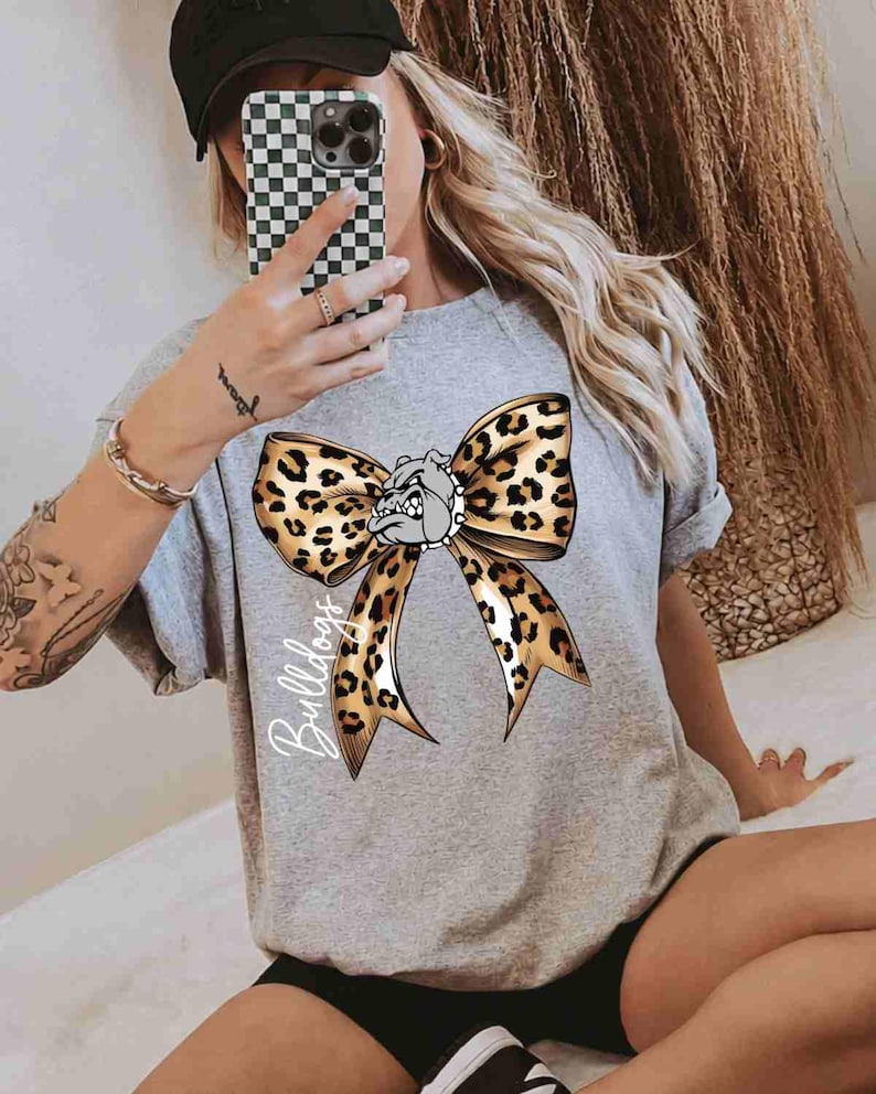 Puede incluir: Una camiseta gris jaspeado de manga corta con un gran estampado gr&aacute;fico de un lazo con estampado de leopardo centrado en el pecho. El lazo incluye una peque&ntilde;a ilustraci&oacute;n de una cara de bulldog en el centro y la palabra Bulldogs escrita en blanco.