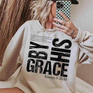 May include: A white sweatshirt with a black graphic print that reads "By His Grace" and "Romans 3: 23-24".