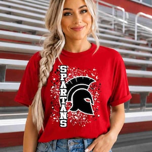 May include: A red short-sleeved t-shirt featuring a black Spartan helmet graphic and the word SPARTANS printed vertically in white block letters, set against a white and black paint splatter design on the front of the garment.