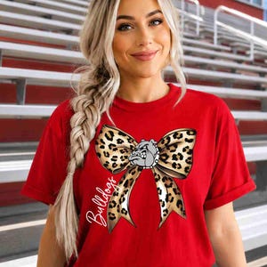 Puede incluir: Una camiseta roja de manga corta con un gran gr&aacute;fico de un lazo con estampado de leopardo centrado en el pecho. El lazo incluye una peque&ntilde;a ilustraci&oacute;n de una cabeza de bulldog en el centro y la palabra Bulldogs escrita en cursiva blanca.