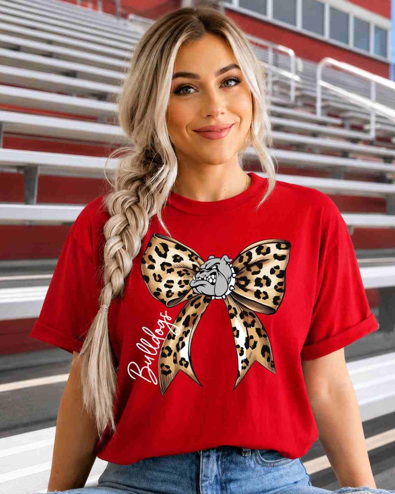 Puede incluir: Una camiseta roja de manga corta con un gran gr&aacute;fico de un lazo con estampado de leopardo centrado en el pecho. El lazo incluye una peque&ntilde;a ilustraci&oacute;n de una cabeza de bulldog en el centro y la palabra Bulldogs escrita en cursiva blanca.