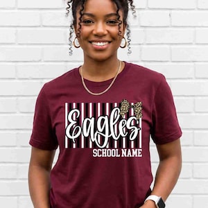 May include: A maroon short-sleeve t-shirt featuring a graphic design with vertical black and white stripes, the word "Eagles" in white script, a leopard print bow, and the text "SCHOOL NAME" in white block letters centered on the chest.
