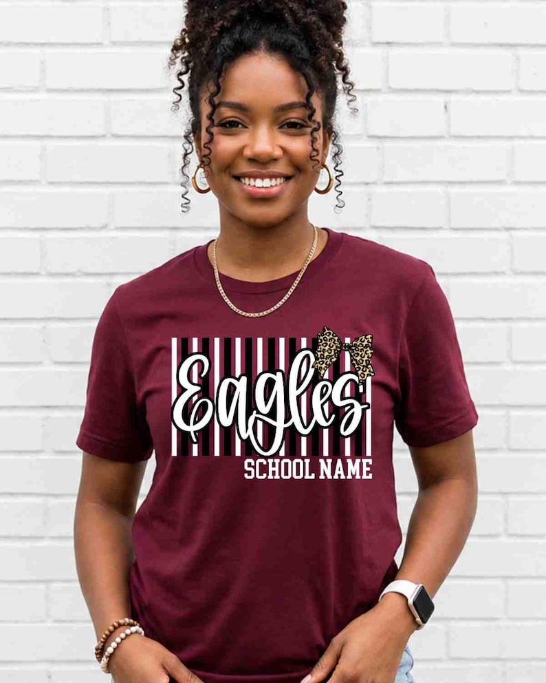 May include: A maroon short-sleeve t-shirt featuring a graphic design with vertical black and white stripes, the word "Eagles" in white script, a leopard print bow, and the text "SCHOOL NAME" in white block letters centered on the chest.