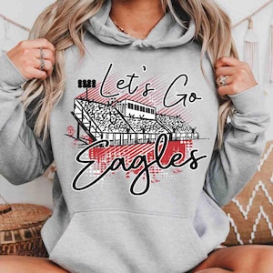 May include: A light gray hoodie with a drawstring hood and a front pocket. The hoodie features a graphic of a stadium with the words "Let's Go Eagles" in black script and red accents.