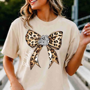 Puede incluir: Una camiseta beige de manga corta con un gr&aacute;fico grande de un lazo con estampado de leopardo. Centrada en el lazo hay una cabeza de bulldog gris con un collar de pinchos. La palabra Bulldogs est&aacute; escrita en cursiva blanca a la izquierda.