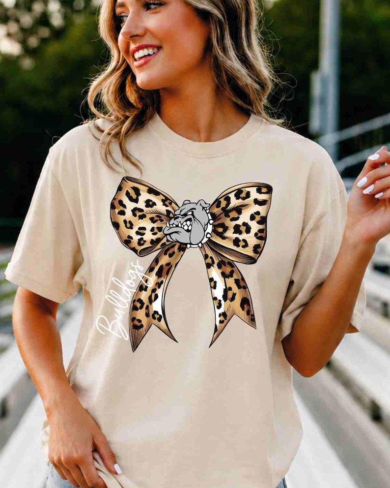 Puede incluir: Una camiseta beige de manga corta con un gr&aacute;fico grande de un lazo con estampado de leopardo. Centrada en el lazo hay una cabeza de bulldog gris con un collar de pinchos. La palabra Bulldogs est&aacute; escrita en cursiva blanca a la izquierda.