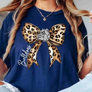 Puede incluir: Una camiseta azul marino con un gr&aacute;fico grande de un lazo con estampado de leopardo centrado en el pecho. Una cabeza de bulldog gris con un collar de pinchos est&aacute; colocada en el centro del lazo. La palabra Bulldogs est&aacute; escrita en cursiva blanca.