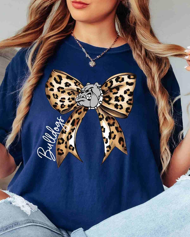Puede incluir: Una camiseta azul marino con un gr&aacute;fico grande de un lazo con estampado de leopardo centrado en el pecho. Una cabeza de bulldog gris con un collar de pinchos est&aacute; colocada en el centro del lazo. La palabra Bulldogs est&aacute; escrita en cursiva blanca.