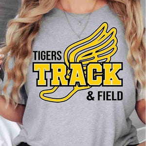 May include: A heather gray short-sleeve t-shirt featuring a graphic design with the text TIGERS TRACK & FIELD in black and yellow lettering. The design includes a large yellow winged emblem positioned behind the word TRACK.