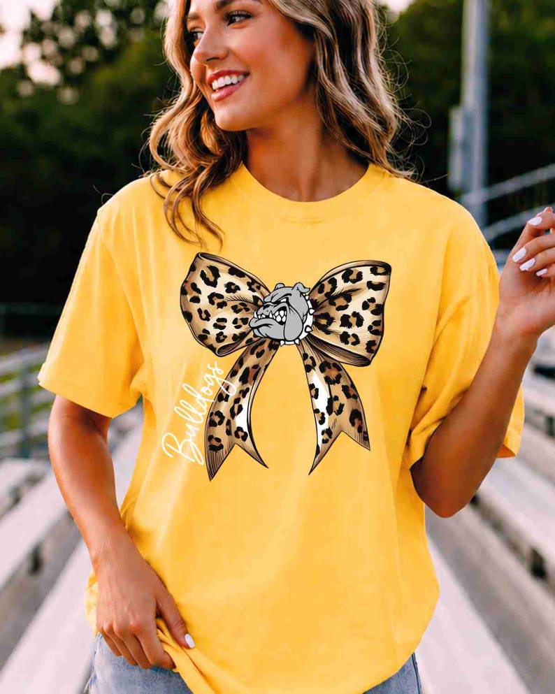 Puede incluir: Una camiseta amarilla de manga corta con un gr&aacute;fico grande de un lazo con estampado de leopardo centrado en el pecho. El lazo incluye una peque&ntilde;a ilustraci&oacute;n de una cara de bulldog gris en el centro y la palabra Bulldogs escrita en blanco.
