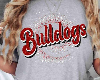 May include: A heather gray short-sleeve t-shirt featuring the word Bulldogs in a red, white, and black script font. The text is layered over a circular graphic of white and red starbursts and sparkles, creating a textured, glittery appearance.