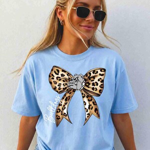 Puede incluir: Una camiseta de manga corta de color azul claro con un gr&aacute;fico grande de un lazo con estampado de leopardo centrado en el pecho. El dise&ntilde;o del lazo incorpora una peque&ntilde;a ilustraci&oacute;n de una cara de bulldog en el centro y la palabra Bulldogs en blanco.