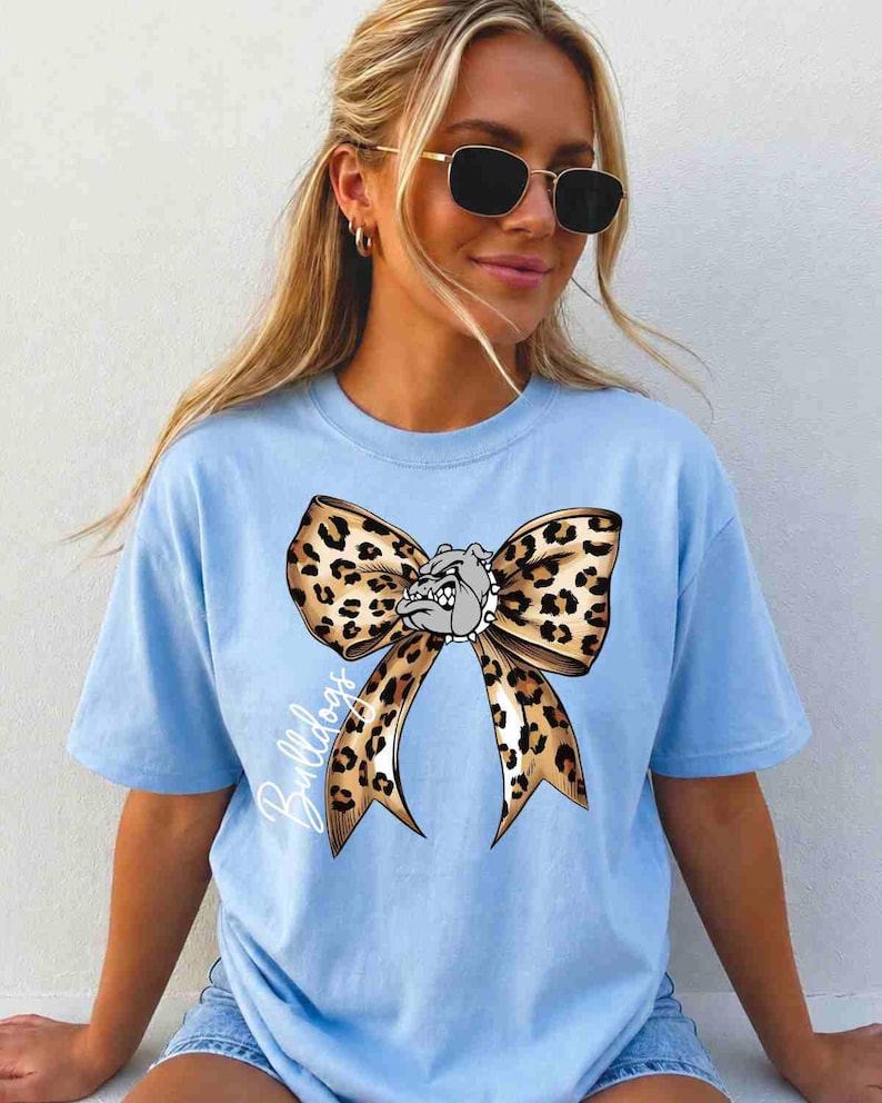 Puede incluir: Una camiseta de manga corta de color azul claro con un gr&aacute;fico grande de un lazo con estampado de leopardo centrado en el pecho. El dise&ntilde;o del lazo incorpora una peque&ntilde;a ilustraci&oacute;n de una cara de bulldog en el centro y la palabra Bulldogs en blanco.