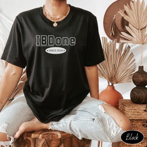 May include: Black t-shirt with the text "IBDone" in white, with "SINCE 2016" below. The shirt is worn by a person wearing gold jewelry. The person is sitting on a wooden stool.