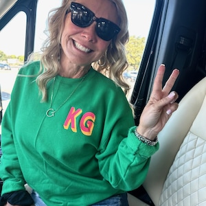 May include: A woman wearing a green sweatshirt with the letters "KG" in pink and yellow on the front. She is wearing denim shorts and sunglasses and is giving a peace sign.