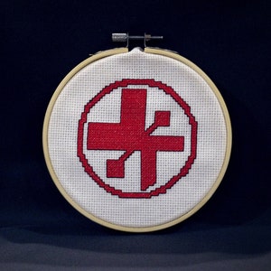 May include: A white embroidery hoop with a red cross and a red symbol inside a circle. The cross and symbol are stitched on a white fabric.