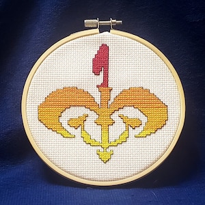 Puede incluir: Un aro de bordado blanco con un diseño de flor de lis bordado en amarillo, naranja y rojo.