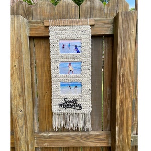 May include: A white woven wall hanging with three photo frames and the words "Sweet Memories" in black lettering. The wall hanging has a fringe at the bottom.