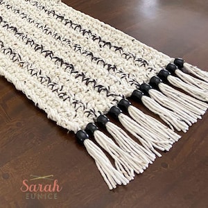May include: Cream-colored, hand-crocheted table runner with dark brown accents. The runner features a textured stitch pattern and is finished with white tassels and black beads. A decorative piece for a dining table or entryway.