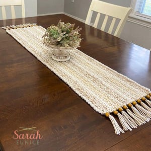 May include: A cream-colored, hand-crocheted table runner with a textured design. The runner has fringe and wooden bead accents at each end. A decorative floral arrangement sits on a pedestal in the center. The table is made of dark wood.