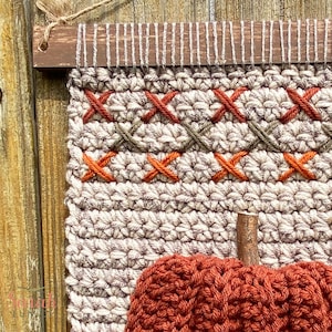 May include: A crocheted wall hanging with a pumpkin design. The hanging features a beige background with orange and green embroidery. The pumpkin is a deep orange color.