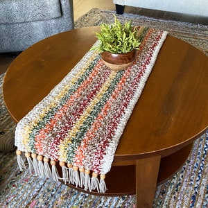 May include: A handmade crochet table runner with a striped pattern in cream, yellow, orange, green, and burgundy. The ends have white fringe with wooden beads. A small wooden bowl with greenery sits on the runner.