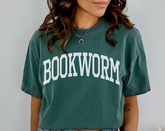 May include: A dark green t-shirt with the word "BOOKWORM" printed in white on the front. The shirt is worn by a person with long brown hair and is paired with light blue denim shorts.