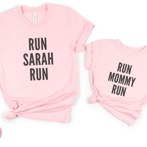 May include: Two pink t-shirts with black text. The larger shirt says "Run Sarah Run". The smaller shirt says "Run Mommy Run".