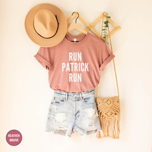 May include: A heather mauve t-shirt with the text "RUN PATRICK RUN" in white lettering. The shirt is hanging on a wooden hanger with a straw hat and a woven bag.