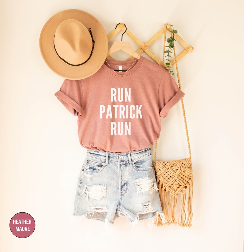 May include: A heather mauve t-shirt with the text "RUN PATRICK RUN" in white lettering. The shirt is hanging on a wooden hanger with a straw hat and a woven bag.