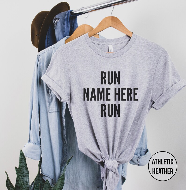 May include: Gray t-shirt with the text "RUN NAME HERE RUN" printed in black. The shirt is on a hanger and has a knotted bottom.