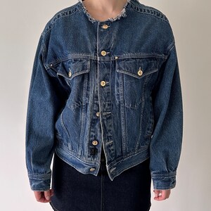 May include: A blue denim jacket with a frayed collar, long sleeves, and multiple pockets. The jacket has a button closure and gold-toned buttons.