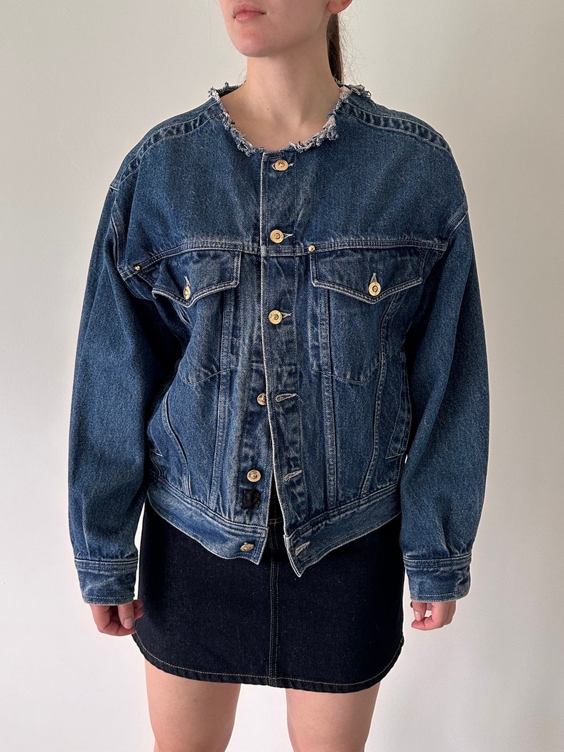 May include: A blue denim jacket with a frayed collar, long sleeves, and multiple pockets. The jacket has a button closure and gold-toned buttons.