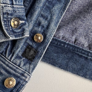 May include: Close-up of a blue denim jacket with gold metal buttons. The buttons have the text "Versace Jeans Couture" engraved on them.